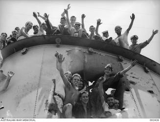 ALEXANDRIA, EGYPT. CREW MEMBERS OF HMAS SYDNEY WAVE GREETINGS FROM THE TOP OF AND A SHELL HOLE IN ONE OF THE SHIP'S FUNNELS. THE SAILOR FIFTH FROM THE LEFT IN THE TOP GROUP IS LEADING STOKER THOMAS NEVIN GLACKIN.